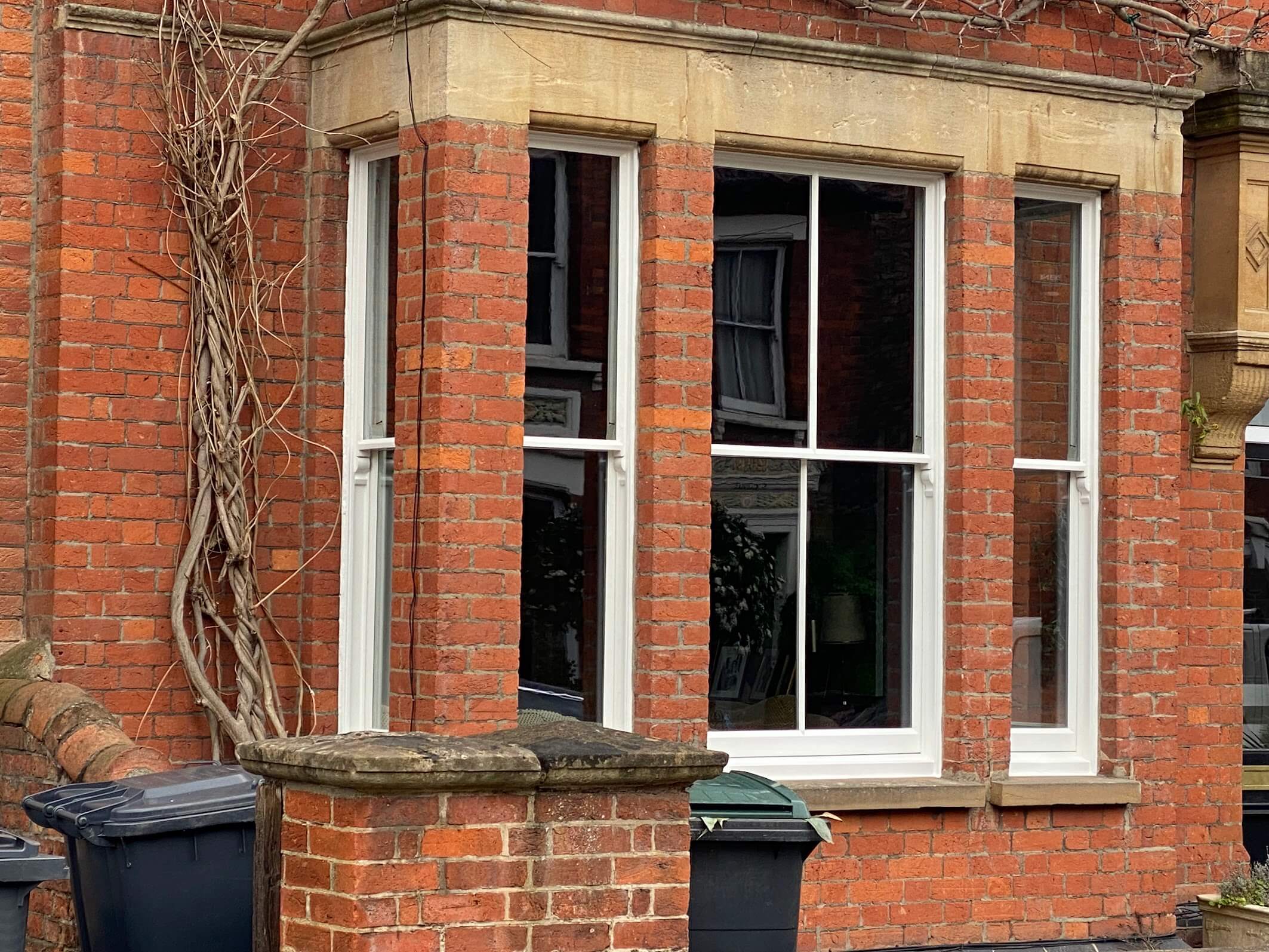 A red brick house with a bay window, white sash windows Cambridge style, a stone window ledge, and a few rubbish bins on the pavement.